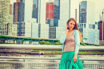 Lonely girl wants someone to talk. Wearing light blue sweater, green skirt, an American woman standing by metal fence on pier in New York, sad, depressed, listening to cell phone. .