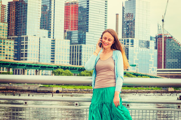 Good to hear from you. Wearing light blue sweater, green skirt, a pretty American woman standing by metal fence on pier in New York, smiling, listening, talking on mobile phone. .