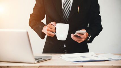 businessman sitting at desk looking at mobile phone holding cup of coffee in office.