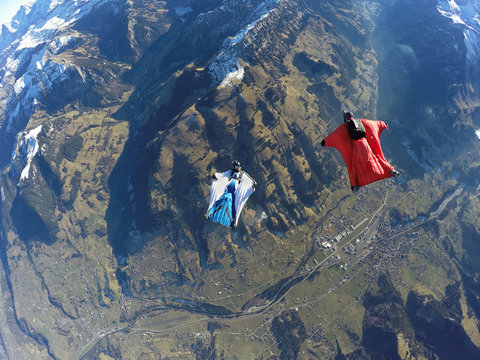 Aerial view of two wingsuit flyers, one on his back facing one in red suit flying above landscape