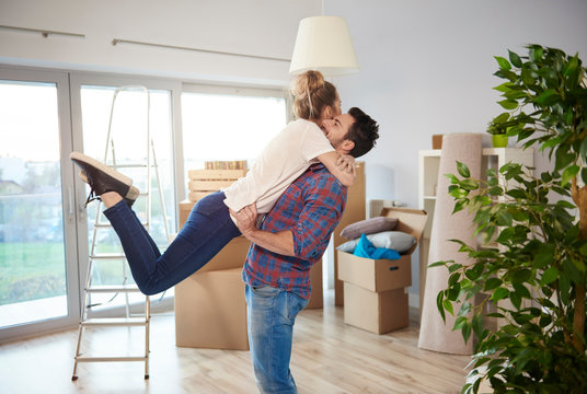 Young couple at home, surrounded by cardboard boxes, man lifting woman in excited hug