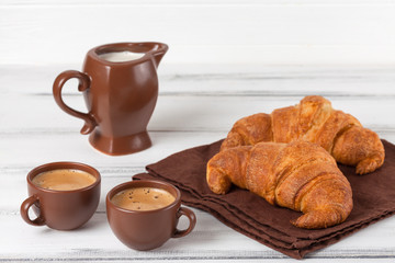 Freshly baked croissants on brown napkin, cream, to cups of coffee in ceramic dishes on white wooden background. Fresh pastries for breakfast. Delicious dessert. Closeup photography.Horizontal banner