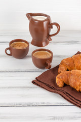 Freshly baked croissants on brown napkin, cream, to cups of coffee in ceramic dishes on white wooden background. Fresh pastries for breakfast. Delicious dessert. Closeup photography. Vertical banner