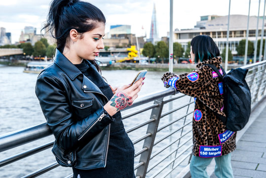 Young woman looking at smartphone with friend looking out from millennium footbridge, London, UK
