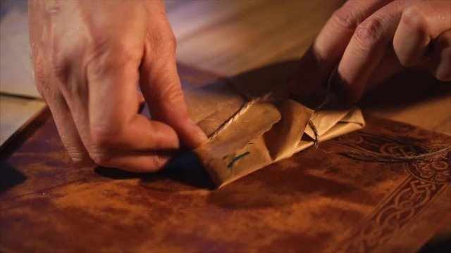 Human is packing little secret letter in a paper and knitting by rope. Close-up of his hands in a table in dark room.