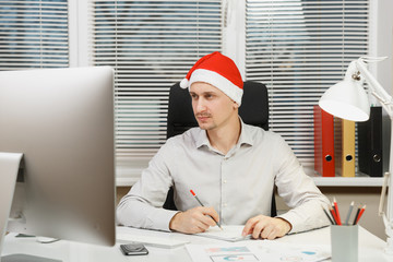 Handsome serious and engrossed business man in shirt, red Christmas hat sitting at the desk,...