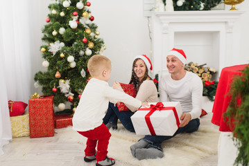 Obraz na płótnie Canvas Happy young cheerful parents with cute little son in red hat. Child boy in light room at home with decorated New Year tree and gift boxes. Christmas good mood. Family, love and holiday 2018 concept.
