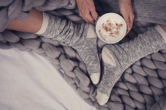 Female feet wearing cozy warm wool socks close up. Woman covered with warm blanket drinking coffee in bed .