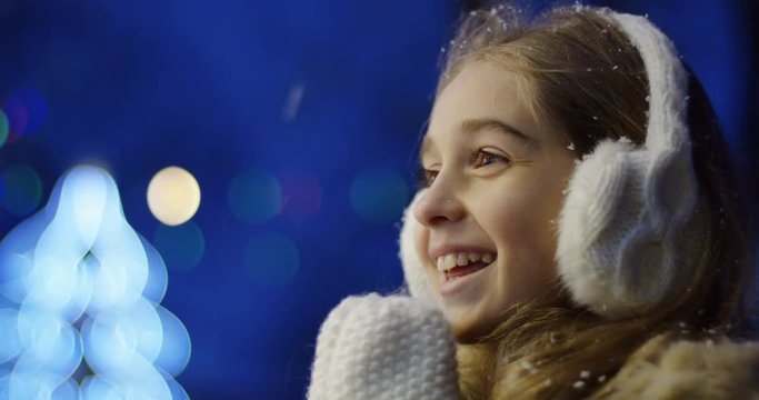 Close up of the happy smiled girl looking at something and clapping in white winter gloves on the blurred Christmas lights in evening. Portrait. Outdoors
