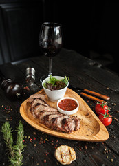 slices of beef on grill with sauce and salad on a wooden plate in serve with a glass of red wine on a dark wooden background - 183857402