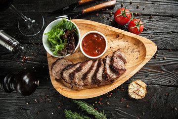 slices of beef on grill with sauce and salad on a wooden plate in serve with a glass of red wine on a dark wooden background - 183857426