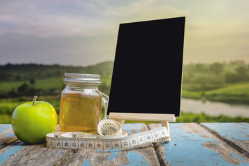 A jar of apple vinegar , measuring tape,  mini blackboard and apple on wooden table in the sunlight with  a blurred nature  background.