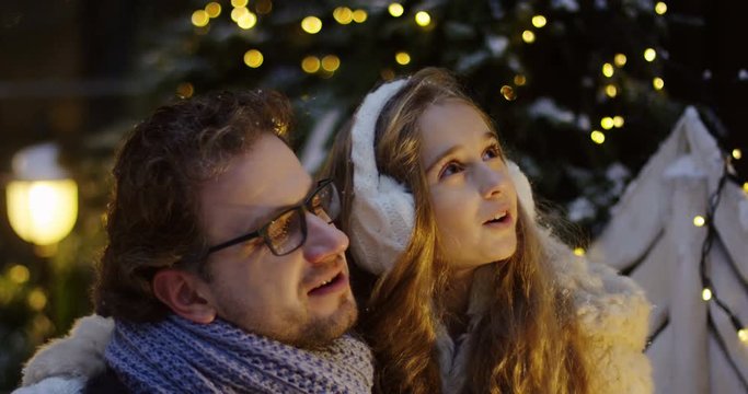 Handsome man in glasses hugging with his pretty daughter, talking and looking fascinated up surrounded by the Christmas decorations on the street in the late evening. Outside. Close up. Portrait shot
