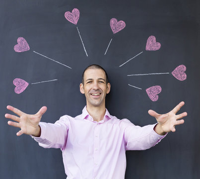 Single adult white man wearing a pink shirt with streched out arms standing in front of a blackboard with painted hearts. Concept of crazy love