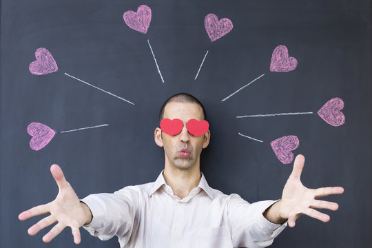 Single adult white man with heart shaped eyes wearing a white shirt and standing in front of a blackboard with painted hearts, stretching out his arms. Concept of crazy love