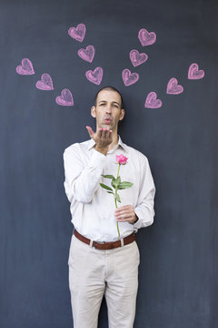 Single adult white man wearing a white shirt standing in front of a blackboard with painted hearts holding a rose. Concept of crazy love