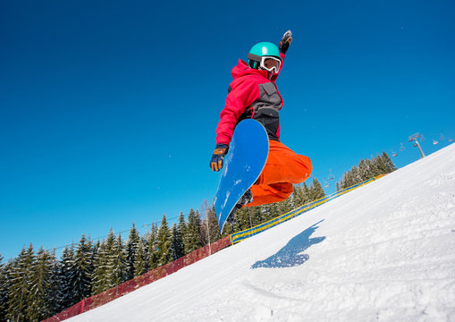 Low angle shot of a snowboarder jumping in the air while riding on the slope in the mountains on a beautiful sunny winter day. Forest, blue sky and ski-lift on the background
