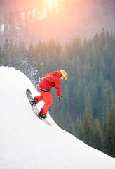 Fototapeta na wymiar Male snowboarder freerider in a red suit riding from the top of the snowy hill with snowboard in the evening at sunset. Skiing and snowboarding concept