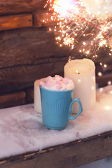 blue mug of drink with whipped cream and marshmallows,candles  on a table covered with snow and sparkler glow