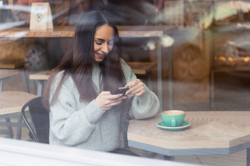 portrait beautiful smiling young woman using mobile phone in cafe throuth glass window