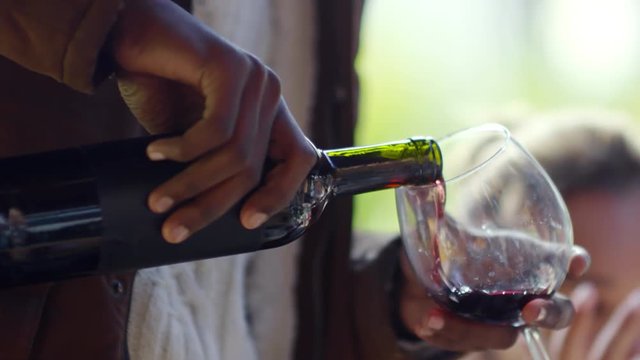 Tilt up of African man smiling and pouring red wine into the glass at outdoor party with friends
