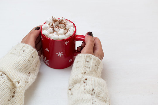 Woman hands in white sweater hold cacao drink,coffee with marshmallow in red mug. White background. Winter style photo.
