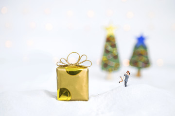 Miniature couple on the white snow with gift box over blurred Christmas tree on white background