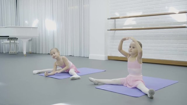 In dance studio two girls sit on mats and learn how to do movements. Little children in skirts attend training on ballet and synchronously repeat exercises.