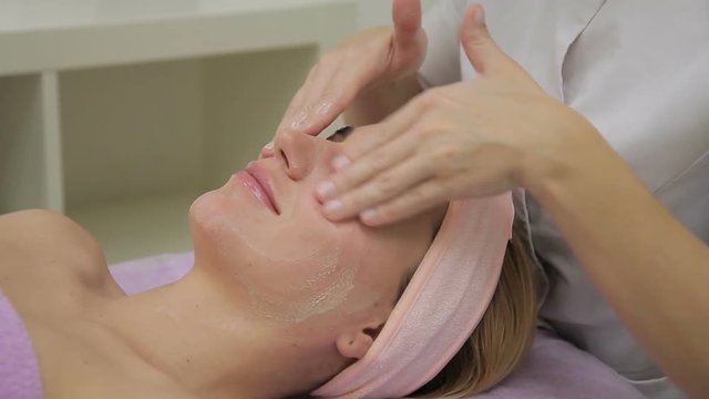 The cosmetologist hands anoint the young female face with hydrating cream in luxury spa salon. The client is laying on the couch covered with perivinkle towel, her dyed blond hair are hold with pink