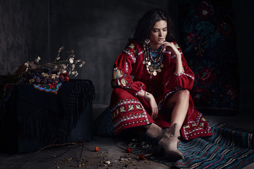 Beautiful young sexy woman with long brown hair, wearing red dress with traditional ornament and light brown boots, sitting on the floor in dark studio