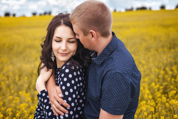 Happy young couple hugging on yellow meadow. Girl with long brunette hair in black dress in white peas, boyfriend in blue shirt in pattern