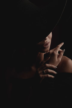 Dramatic dark studio portrait of elegant and luxury woman with beautiful hands, wearing black wide hat and black dress. Hidden eyes.