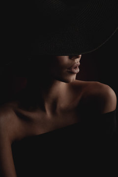 Dramatic dark studio portrait of elegant and luxury woman with beautiful hands, wearing black wide hat and black dress. Hidden eyes.