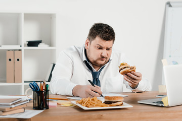 overweight businessman working while eating hamburger and french fries in office