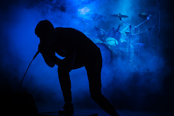 Silhouette of a singer with a microphone at a concert on the background of drums in smoke with blue lighting