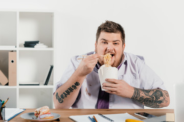 overweight tattooed businessman eating noodles at workplace in office