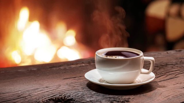 Cup of coffee stands on an old wooden board in front of a burning fireplace. Steam rises from a cup of coffee. The looped video of 1920x1080