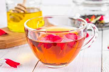 A cup of tea, in the background of a bank of honey and a jar with a black herbal floral tea on a white wooden table.