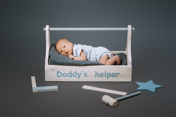little baby in bodysuit lying in wooden toolkit with daddys helper lettering and hammer near by