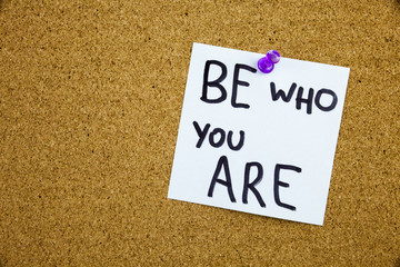 A yellow sticky note writing, caption, inscription chalkboard with the phrase be who are you in black ext on a sticky note pinned to a cork notice board