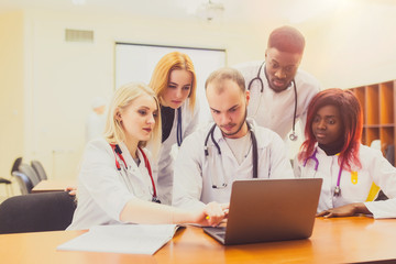 A group of mixed-race physicians discusses the diagnosis behind a laptop in the office. In the background, the asian professor explains to students the physicians anatomy