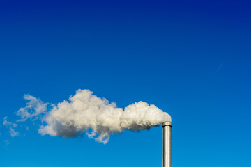 A metallic chimney giving off a heavy cloud of white smoke against a deep blue sky.