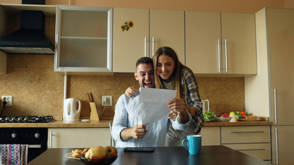 Attractive happy couple recieve good news unfolding letter in the kitchen while have breakfast at...