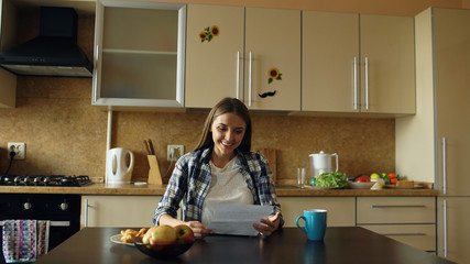 Attractive happy woman recieve good news reading letter in the kitchen while have breakfast at home...