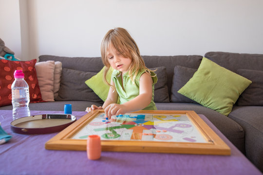 four years old blonde child with green sleeveless shirt sitting on brown sofa, playing parcheesi or parchisi or parchis, on table
