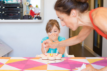 Obraz na płótnie Canvas woman putting candles in birthday cake on colorful tablecloth at home and three years old blonde child with expression of emotion 