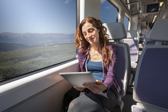 Red Hair Smiling Woman Dressed In Purple And Blue, Traveling By Train Sitting Reading Digital Tablet Or Ebook
