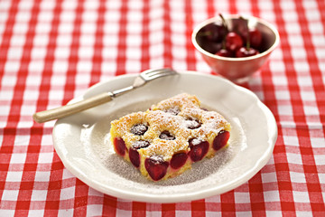 Ein Stück Kirschkuchen auf einer rot-karierten Tischdecke und eine Schale mit frischen Kirschen
