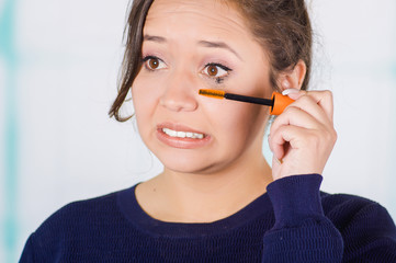 Close up of worried young woman doing a mess using a eye mascara in her eye, in a blurred background