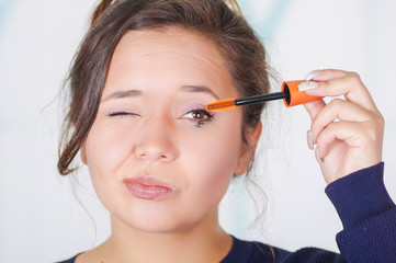 Close up of worried young woman, closing her eye while she is doing a mess using a eye mascara in her eye, in a blurred background